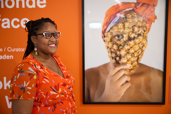 Une femme souriante debout à côté d’une photo d’une personne avec un foulard sur la tête et une partie d’un ananas couvrant son visage et montrant juste ses yeux.