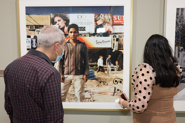 Un homme et une femme vus par derrière regardant une photo d’un homme debout devant une scène de marché avec des panneaux d’affichage et des gens en toile de fond.