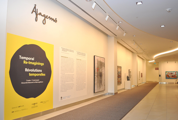 Un espace de galerie avec « Âjagemô » écrit en cursive au-dessus d’un grand graphique et des panneaux de texte et des œuvres d’art installées sur les murs plus bas dans l’espace.