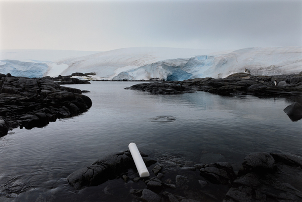 Un tube cylindrique blanc repose sur une surface rocheuse au bord de l'eau. Des icebergs sont en arrière-plan.