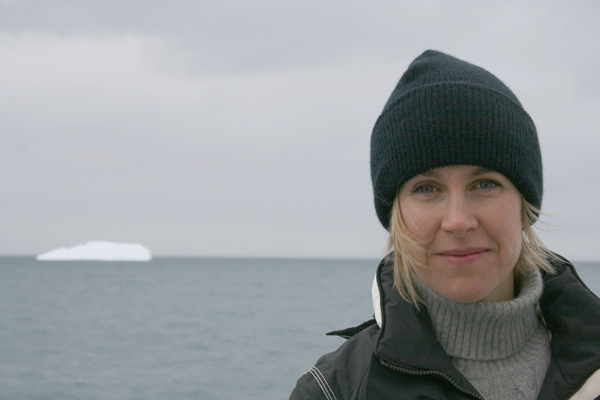 Une femme aux cheveux blonds de la longueur du menton et portant une tuque se tient au premier plan et, par-dessus son épaule droite, un iceberg flotte dans une étendue d'eau.