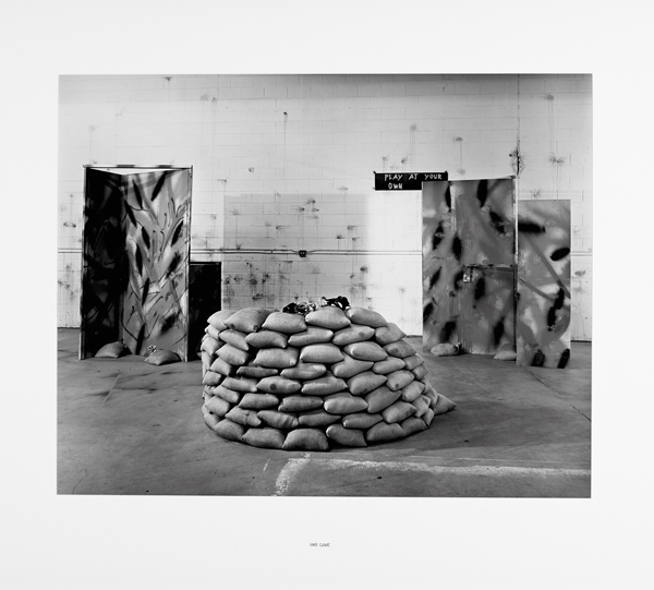 Photo en noir et blanc d'un monticule de sacs de sable placé devant plusieurs panneaux camouflés. Le mur blanc à l'arrière-plan est criblé de traces de balles noires.