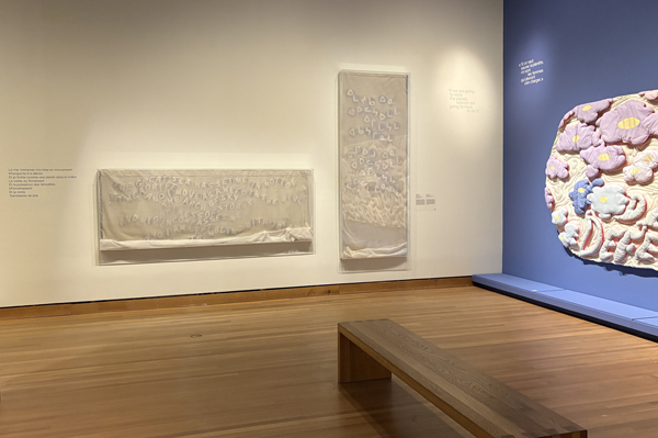 À gauche, une œuvre d'art textile pâle dont les lettres sont recouvertes d'un tissu transparent est suspendue horizontalement dans un boîtier en plexi, à côté d'une autre œuvre suspendue verticalement. À droite, une grande œuvre textile colorée avec des mots gonflés et des fleurs est accrochée à un mur bleu.