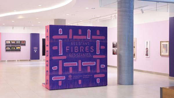 Une exposition intitulée « Resistant Fibres / Fibres Résistantes » présente une grande installation violette représentant le titre et diverses œuvres d'art textile, dont un sac à feu métis orné de perles accroché au mur.
