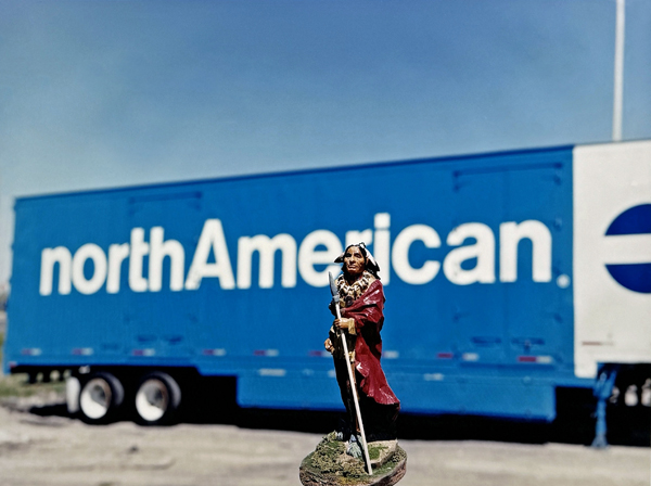 À l'arrière-plan se trouve une remorque de tracteur bleue sur laquelle est inscrit « northAmerican. ». Au premier plan, une figurine en plastique représentant un chef autochtone.
